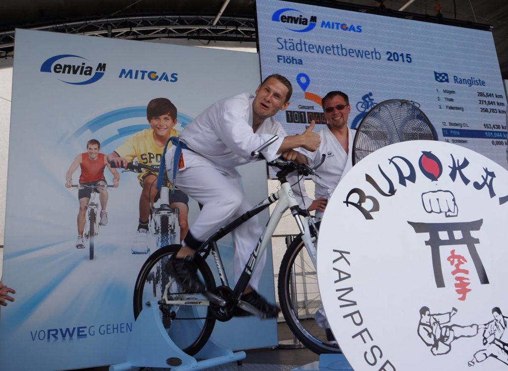 Foto mit zwei Kampfsporterln auf der Bühne. Einer auf dem Fahrrad mit Daumen nach oben. Dahinter die aktuelle Rangliste und im Vordergrund das Logo des Budokan Kampfsportvereins Flöha